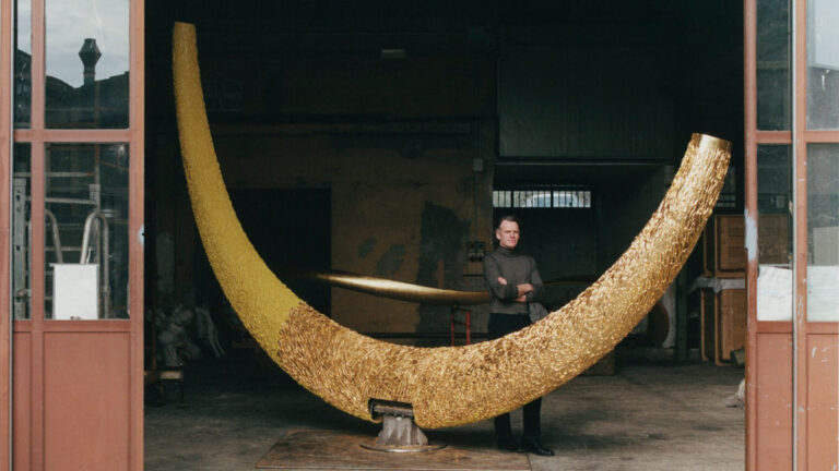 Artist and designer Joseph Walsh stands next to a very large curved object in the open door of his studio