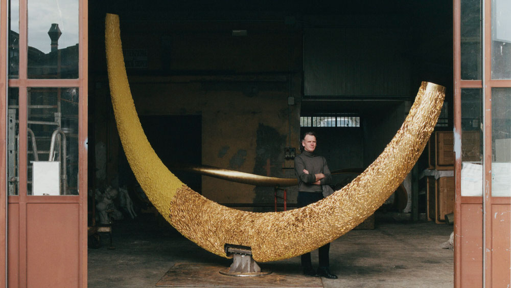 Artist and designer Joseph Walsh stands next to a very large curved object in the open door of his studio