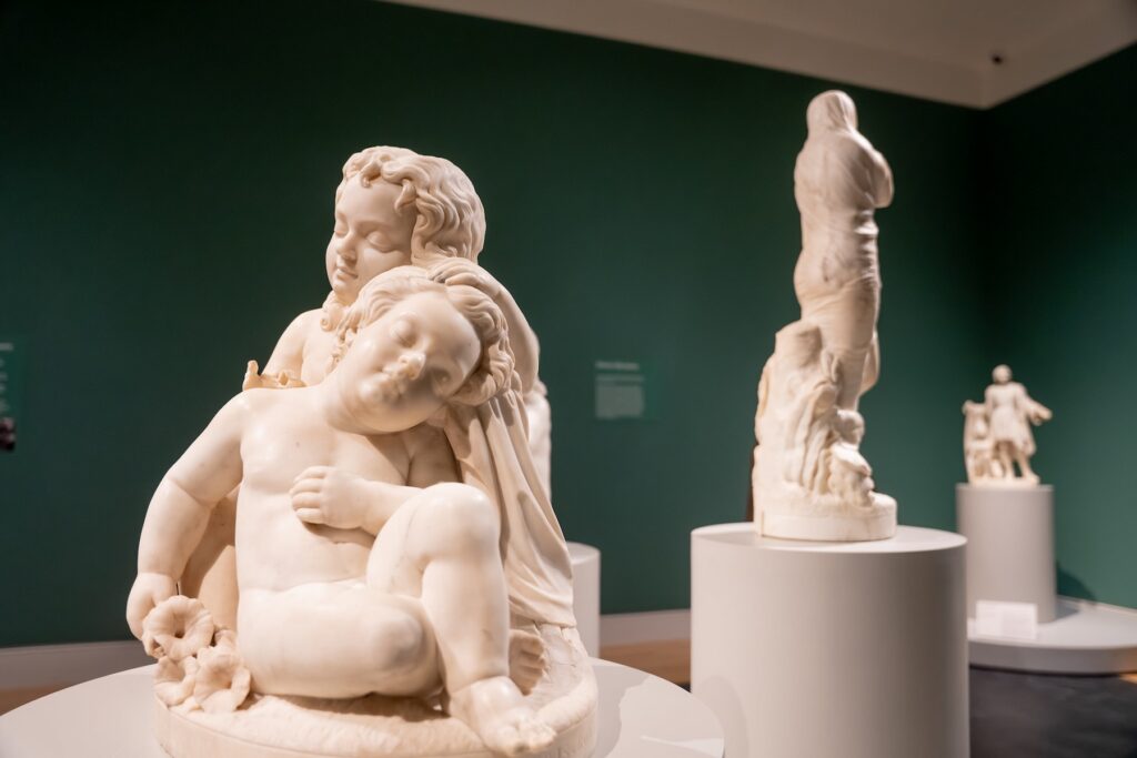 A view of a gallery at the Georgia Museum of Art featuring three of Edmonia Lewis' sculptures. In the foreground, the sculpture is of a young child resting in the arms of another young child. Both of their eyes are closed.