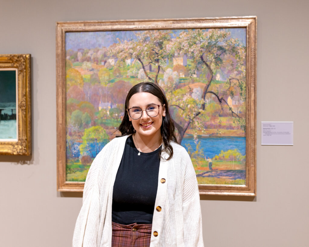 Gallery guide Sarah Gattis stands in the museum's galleries in front of a large painting.