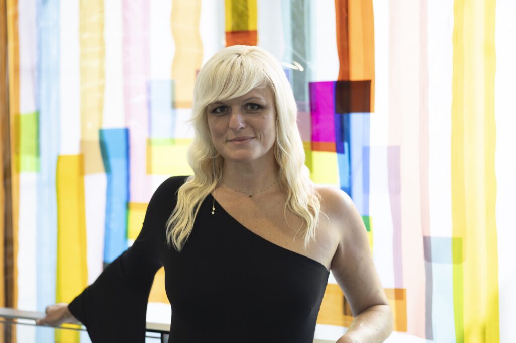 Museum Shop associate Lily Herne leans against a railing in front of a translucent quilt-like artwork by Rachel Hayes.