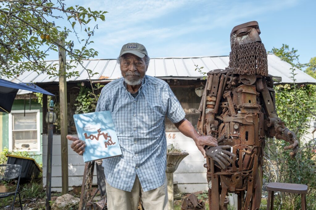 Sculptor Harold Rittenberry stands next to "Byzantine Knight," a large rust-colored metal sculpture welded from repurposed objects. Harold holds a copy of "Harold Rittenberry: Love is All" in his right hand and rests his left hand on the wrist of the "Byzantine Knight."