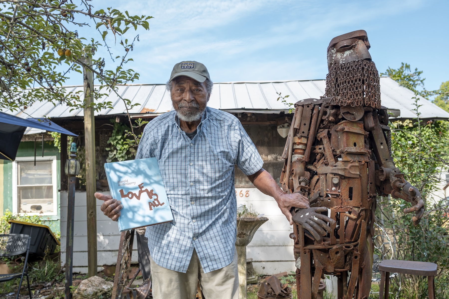 Sculptor Harold Rittenberry stands next to "Byzantine Knight," a large rust-colored metal sculpture welded from repurposed objects. Harold holds a copy of "Harold Rittenberry: Love is All" in his right hand and rests his left hand on the wrist of the "Byzantine Knight."