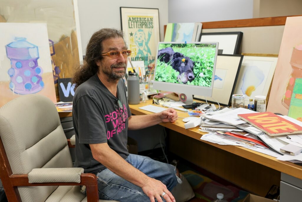 Art handler Larry Forte sits at his desk in front of a computer. Various paintings and prints can be seen in the background.