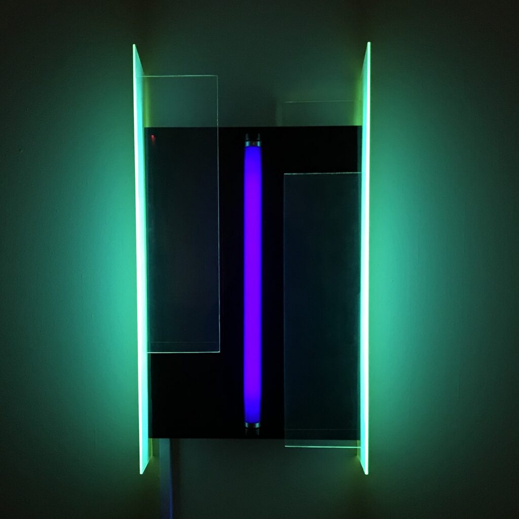 A light sculpture by Lila Katzen that features an ultraviolet light tube installed vertically between two sheets of acrylic. When plugged in and turned on, the acrylic appears to light up too, in two bright vertical bands of light green.