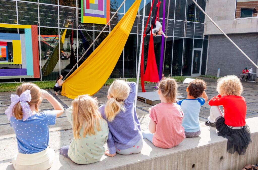 A row of six small children sit on the wall in the museum's sculpture garden to watch an aerial artist perform on yellow and red silks. In the background, two multi-colored works by Rachel Hayes are hanging on the museum's window.