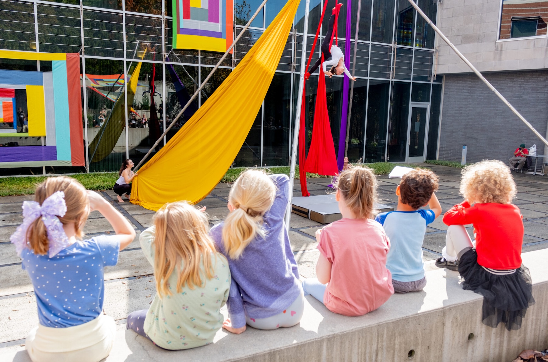 A row of six small children sit on the wall in the museum's sculpture garden to watch an aerial artist perform on yellow and red silks. In the background, two multi-colored works by Rachel Hayes are hanging on the museum's window.
